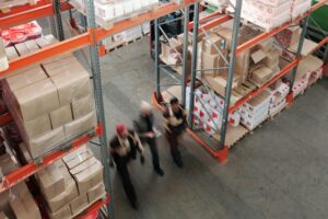 Three warehouse workers walking through a storage area filled with stacked boxes and products on industrial shelving.