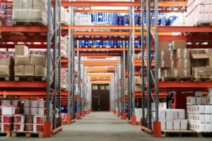 ide-angle view of a well-orwganized warehouse with tall, orange shelving units stacked with various goods, including boxes, containers, and drums. The orderly layout highlights efficient use of space, which is key for maximizing productivity and improving inventory management in a warehouse setting.