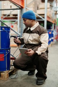 A warehouse worker using a barcode scanner and tablet to manage inventory near a blue barrel