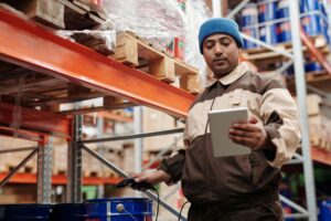 Close-up of a warehouse worker scanning a barcode on a shelf label with a handheld RFID scanner, surrounded by organized shelving units filled with inventory. The image emphasizes the importance of accurate labeling and RFID systems in streamlining warehouse operations and improving efficiency.